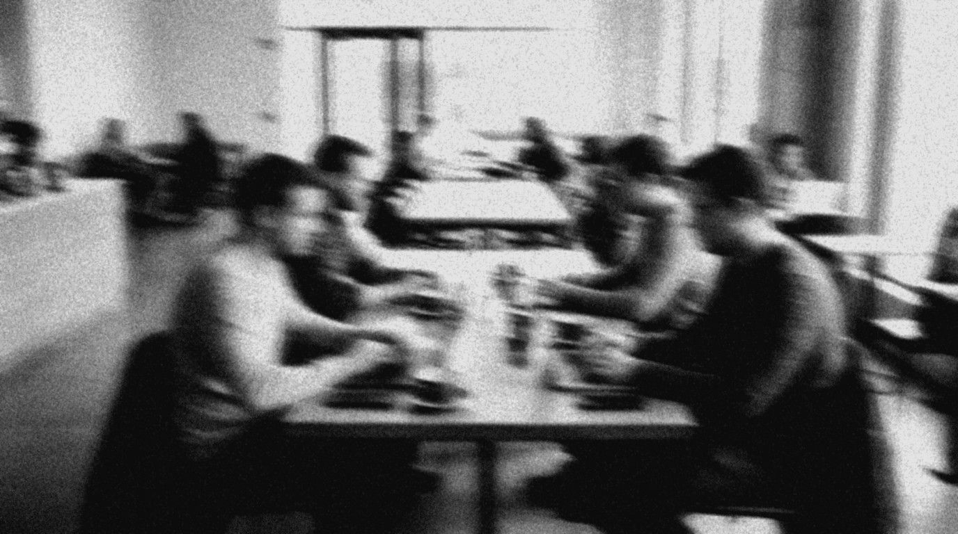 Blurred black-and-white photo: a group working on laptops at a long table in a bright café during the day