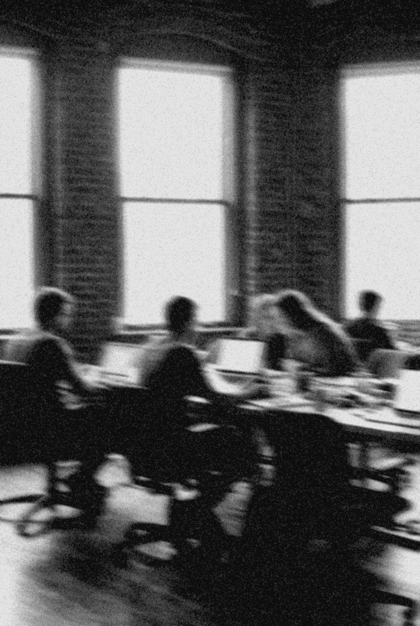 Blurred black-and-white photo: a group at laptops in a bright loft, two people leaning toward each other